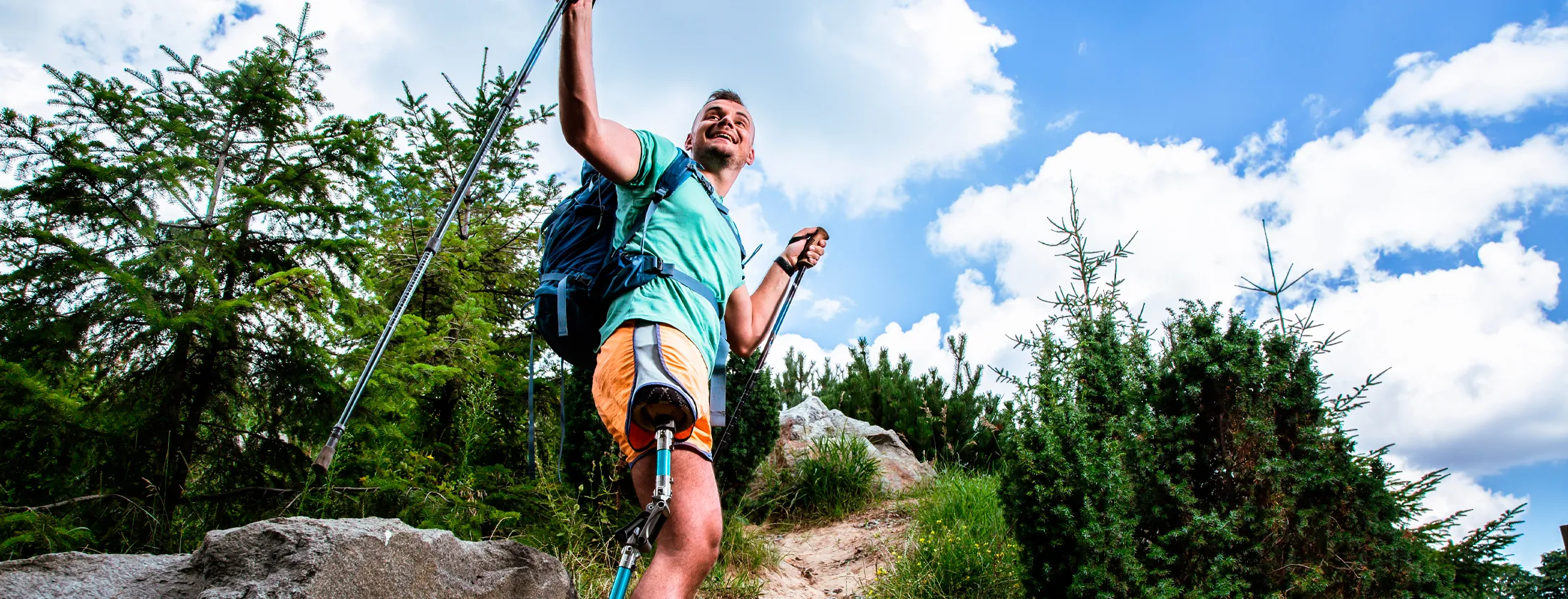 Man with a prosthetic leg hiking seen from a low angle, evergreen trees, trail, and blue sky with clouds in the background